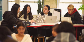 Four students with laptops sit around a table in conversation.