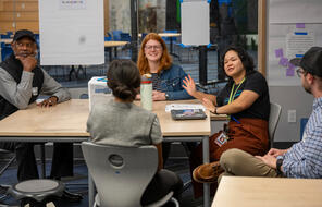 A diverse group of students and teachers discuss something at a table