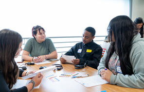 students gathering around a table that has headphones, papers, and multi colored stickers on top of it