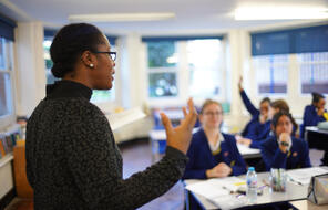 woman wearing a black shirt and wearing glasses standing in front of classroom speaking to students 