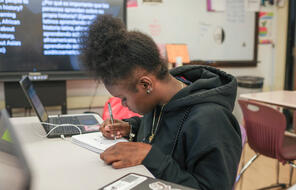 Student wearing a black hoodie writing while sitting at a classroom desk. 