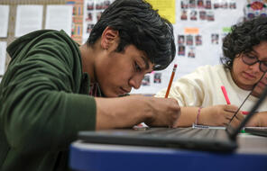 Two students at a desk in a classroom and writing on a piece of paper on their desks