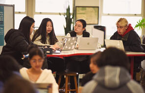 Four students with laptops sit around a table in conversation.