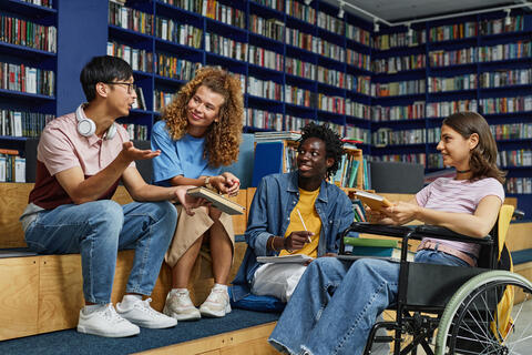 Diverse group of students in college library including young woman in wheelchair enjoying discussion.